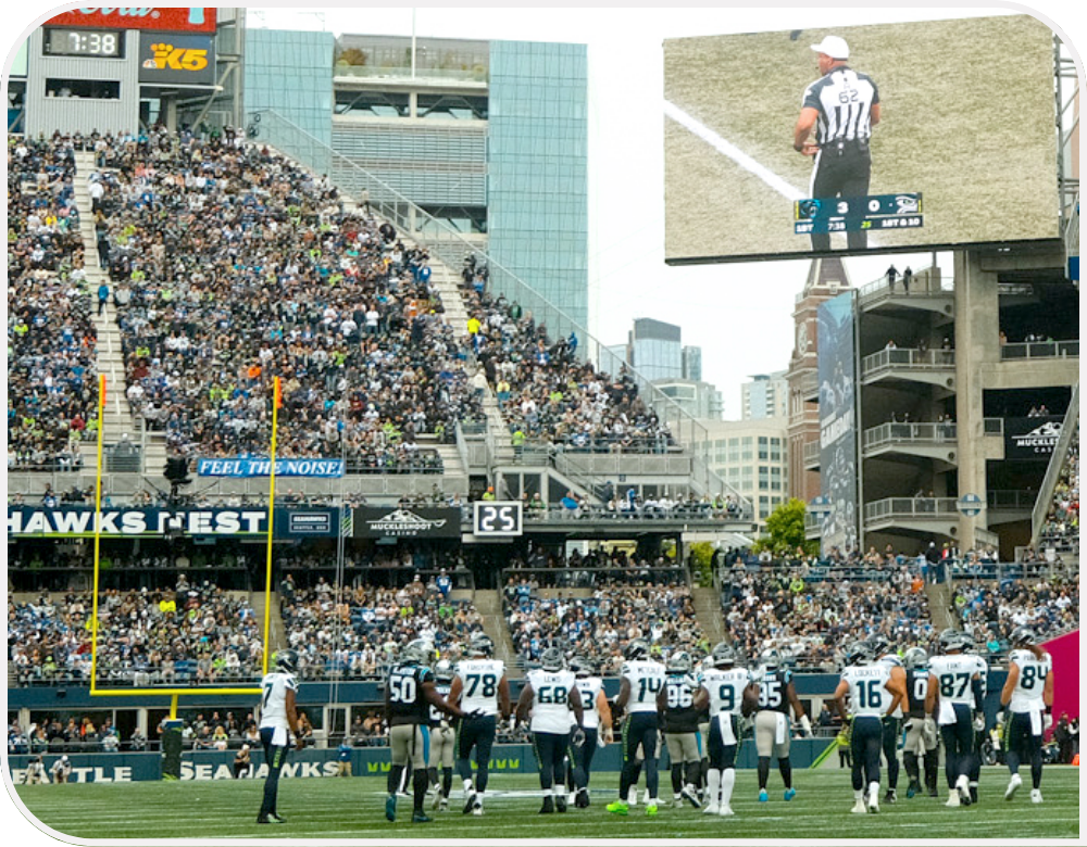 Seahawks on the field at Lumen Field stadium with fans in the background and a referee on the stadium screen.