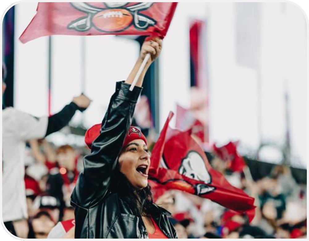 A fan in Buccaneers attire cheers in the stands and waves a team flag. 