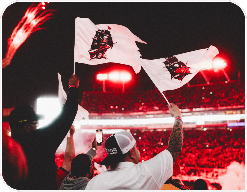 Fans cheer from the stands and wave Buccaneers themed flags excitedly.  
